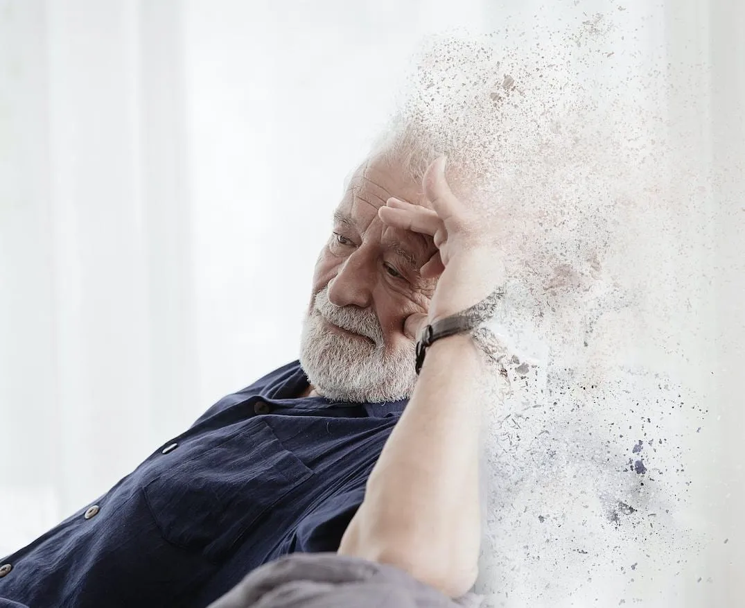 Un homme âgé assis près d’une fenêtre, la tête appuyée sur sa main ; le contour de sa silhouette se dissout en fines particules.